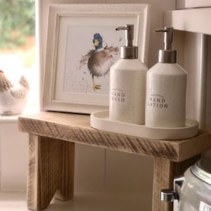 May include: Two off-white speckled ceramic pump bottles labelled Hand Wash and Hand Lotion, resting on an oval tray. Behind them is a framed watercolour print of a mallard duck, all set upon a small, rectangular, light brown wooden riser.