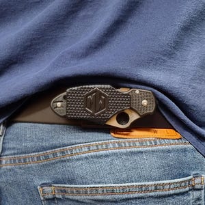 Könnte beinhalten: Ein schwarzes und hellbraunes Taschenmesser mit strukturiertem Griff ist an einem braunen Ledergürtel befestigt. Das Messer hat ein geometrisches Muster und die Initialen "MA". Das Messer wird teilweise von einem blauen Hemd und einer blauen Jeans verdeckt.