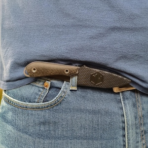 Könnte beinhalten: Ein Messer mit dunklem Griff und strukturierter Klinge steckt in der Tasche einer blauen Jeans. Der Messergriff hat zwei sichtbare Nieten. Die Jeans ist hellblau gewaschen, und das Hemd ist einfarbig blau.