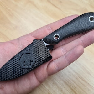 Könnte beinhalten: Ein kleines, schwarzes Messer mit strukturiertem Griff und Scheide. Das Messer hat einen silbernen Ring am Ende des Griffs und zwei silberne Nieten. Die Scheide hat ein geometrisches Muster und ein Logo.