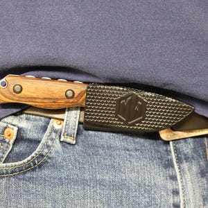 Puede incluir: Un cuchillo con mango de madera y funda negra se guarda en un cintur&oacute;n, visible contra una camisa azul y unos vaqueros. El mango del cuchillo es marr&oacute;n con dos remaches met&aacute;licos. La funda tiene un patr&oacute;n texturizado y un logotipo.