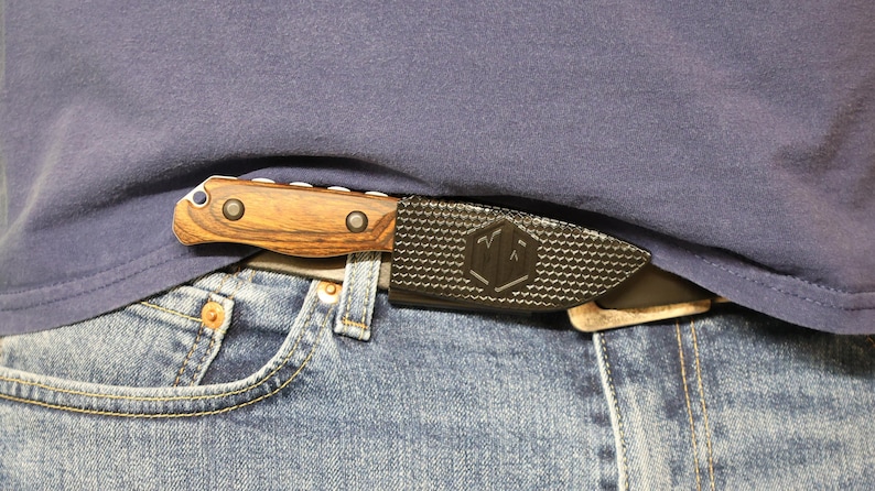 Puede incluir: Un cuchillo con mango de madera y funda negra se guarda en un cintur&oacute;n, visible contra una camisa azul y unos vaqueros. El mango del cuchillo es marr&oacute;n con dos remaches met&aacute;licos. La funda tiene un patr&oacute;n texturizado y un logotipo.