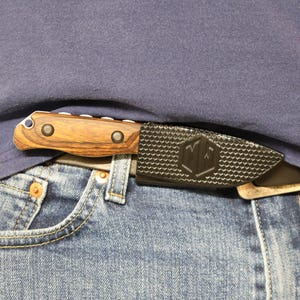 Puede incluir: Un cuchillo peque&ntilde;o de hoja fija con mango de madera y funda negra texturizada. El cuchillo est&aacute; parcialmente oculto, sujeto a un cintur&oacute;n y se lleva con vaqueros azules y una camisa azul. La funda tiene un logotipo.