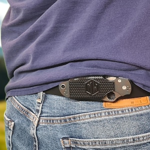 May include: A black and silver folding knife clipped to a black belt. The knife has a textured handle with a geometric pattern and a silver blade. The belt is worn with blue jeans and a blue shirt.
