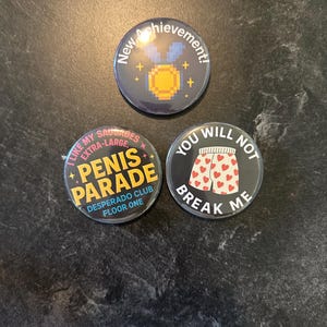 May include: Three round pinback buttons with humorous text. One button says "New Achievement!" with a pixelated gold medal. Another says "I like my sausages extra-large. Penis Parade Desperado Club Floor One." The third says "You will not break me."