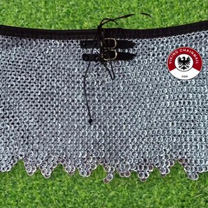 Puede incluir: Una falda de cota de malla plateada con cinturilla negra y correas de cuero con hebillas. La falda tiene un borde inferior festoneado y un logotipo circular que dice "VIKING CHAINMAIL USA".