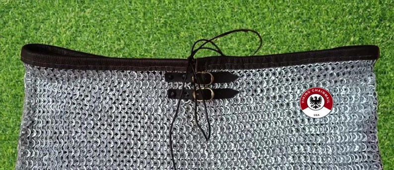 Puede incluir: Primer plano de una prenda de cota de malla con ribete negro y correas de cuero con hebillas. La cota de malla est&aacute; hecha de peque&ntilde;os anillos met&aacute;licos entrelazados. La prenda tiene una etiqueta con un emblema rojo, blanco y negro y el texto "VIKING CHAINMAIL USA".