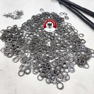 May include: A large pile of silver metal rings, likely for chainmail, with a small pile of metal rivets in the background. The rings are all the same size and shape, and they are arranged in a random pattern. The image also includes a small sticker with the text "Viking Chainmail USA".
