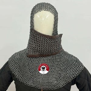 Puede incluir: Cota de malla con capucha y protección para el cuello. La cota de malla está hecha de pequeños anillos metálicos entrelazados. La protección del cuello tiene un ribete de cuero marrón. Un logotipo circular con el texto "VIKING CHAINMAIL USA" es visible.