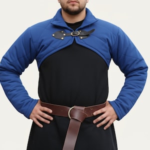 Könnte beinhalten: Ein blaues, gepolstertes Gambeson mit langen Ärmeln und einem Steppmuster. Das Kleidungsstück hat einen schwarzen Lederverschluss mit einer Messingschnalle. Die Person trägt einen braunen Ledergürtel mit einer silbernen Schnalle und ein schwarzes Hemd.