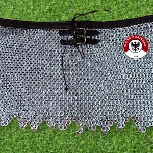 Puede incluir: Una falda de cota de malla plateada con cinturilla negra y correas de cuero con hebillas. La falda tiene un borde inferior festoneado y un logotipo circular que dice "VIKING CHAINMAIL USA".