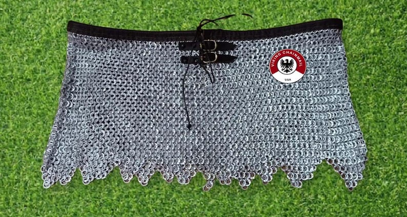 Puede incluir: Una falda de cota de malla plateada con cinturilla negra y correas de cuero con hebillas. La falda tiene un borde inferior festoneado y un logotipo circular que dice "VIKING CHAINMAIL USA".