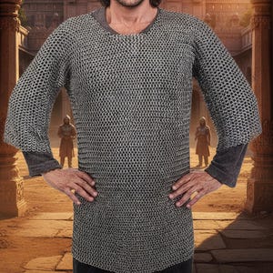 Op de afbeelding: Een zilveren maliënkolder met lange mouwen en een ronde hals. Het shirt wordt gedragen over een grijs shirt met lange mouwen. De maliënkolder is gemaakt van kleine, in elkaar grijpende metalen ringen.