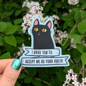 May include: A die-cut sticker featuring a black cat with large yellow eyes and pink inner ears. Below the cat is a light blue banner with the black text: I URGE YOU TO ACCEPT ME AS YOUR RULER. The sticker has a light blue border and matte finish.