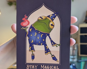 May include: A whimsical illustration of a frog wizard holding a flaming torch, dressed in a blue robe adorned with moons and stars. The image is set against a cream background within a dark blue frame, with the words "STAY MAGICAL" at the bottom.