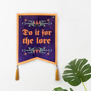 Peut inclure: Une bannière violette avec une bordure orange et le texte "Do it for the lore" en lettres orange. La bannière comporte des éléments floraux décoratifs et des glands dorés en bas.