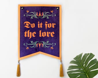 Puede incluir: Un estandarte de pared rectangular de color morado con un borde naranja y dos borlas doradas en la parte inferior. El texto "Do it for the lore" está impreso en naranja en el centro, flanqueado por motivos florales decorativos.