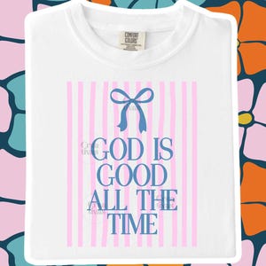 Puede incluir: Una camiseta blanca con un diseño gráfico de rayas verticales de color rosa claro y un lazo azul en la parte superior. El texto "GOD IS GOOD ALL THE TIME" está impreso en letras mayúsculas azules centradas bajo el lazo. Cuello redondo.