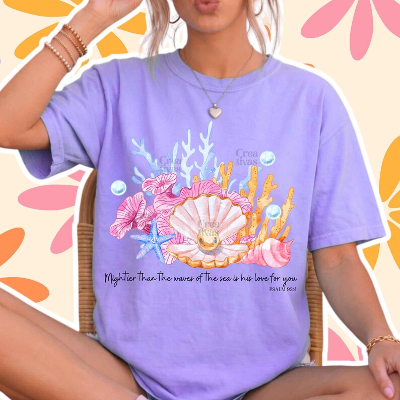 May include: A light purple t-shirt featuring a graphic print of a seashell with a pearl, coral, and a blue starfish. The text reads: Mightier Than The waves of The sea is his love for you PSALM 93:4. The design includes small white bubbles.