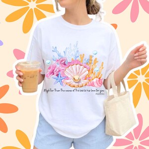 May include: A white t-shirt featuring a colorful graphic of a seashell with a pearl, blue starfish, coral, and bubbles. The text reads, Mightier Than The waves of The sea is his love for you PSALM 93:4. The shirt has a crew neck and short sleeves.
