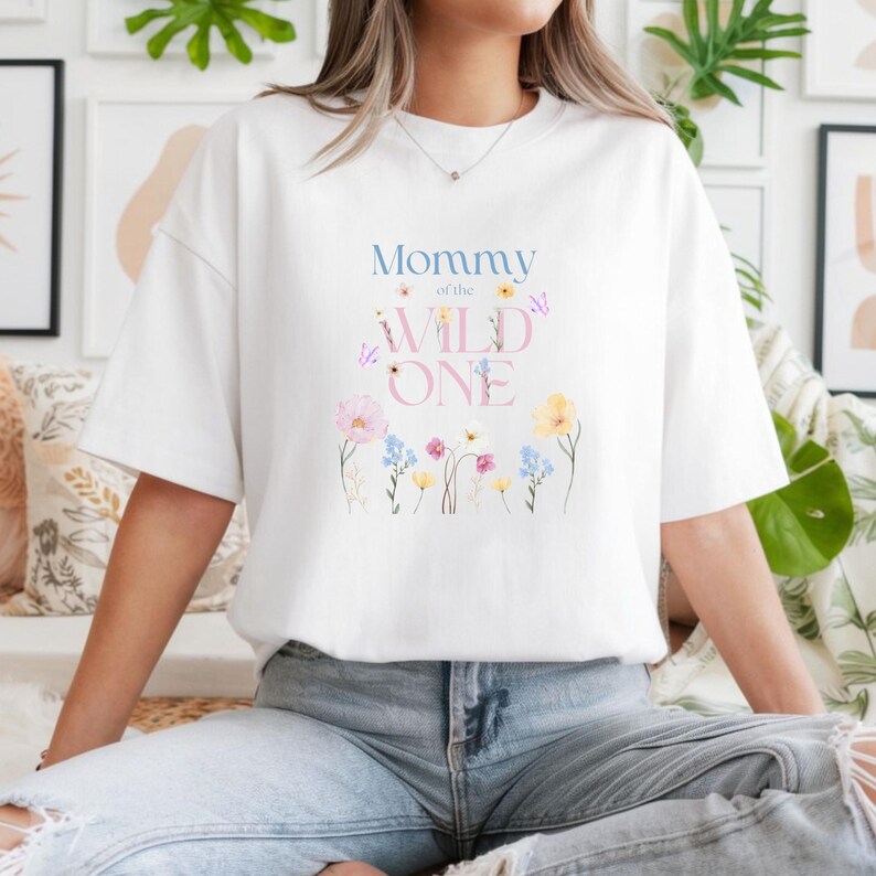 May include: White t-shirt featuring the text "Mommy of the Wild One" in blue and pink, with floral and butterfly illustrations. The shirt has a relaxed fit.