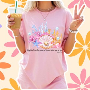 May include: A light pink t-shirt featuring a graphic print of a seashell with a pearl, coral, and a blue starfish. The text "Mightier than the waves of the sea is his love for you" and "PSALM 93:4" is printed in black below the aquatic illustration.