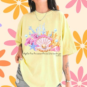 May include: A pale yellow t-shirt featuring a graphic of a pearl inside an open seashell, surrounded by pink and blue coral, a blue starfish, and bubbles. The text reads: Mightier Than The waves of The sea is his love for you PSALM 93:4.