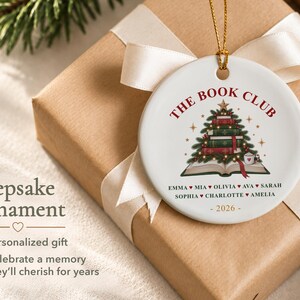 May include: A round white ceramic ornament featuring a graphic of a Christmas tree made of stacked books atop an open book. The text reads THE BOOK CLUB, followed by a list of names separated by hearts and the year 2026 in gold at the bottom.