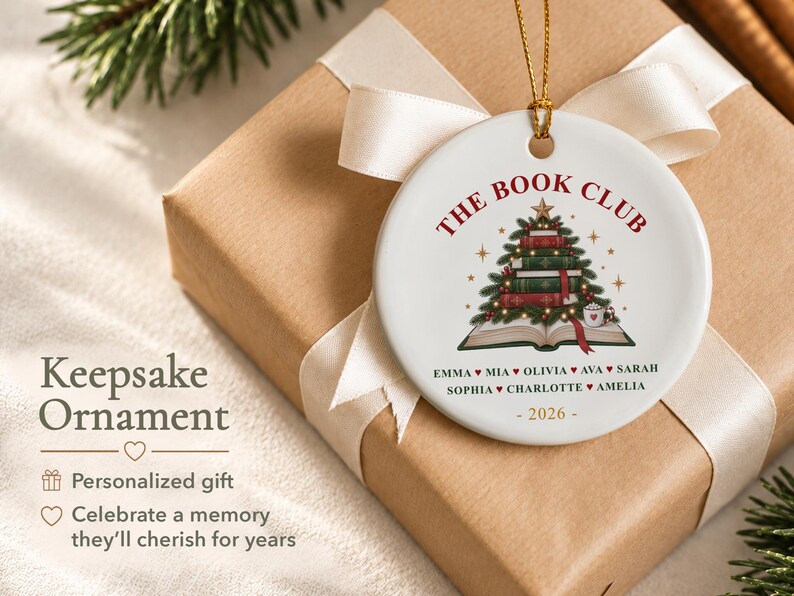 May include: A round white ceramic ornament featuring a graphic of a Christmas tree made of stacked books atop an open book. The text reads THE BOOK CLUB, followed by a list of names separated by hearts and the year 2026 in gold at the bottom.
