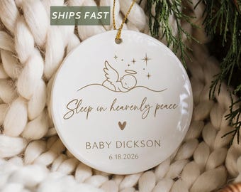 May include: A round white ceramic ornament featuring a gold line drawing of a sleeping angel with a halo and stars above. The text reads Sleep in heavenly peace, BABY DICKSON, and 6.18.2026, with a small gold heart centered below the text.