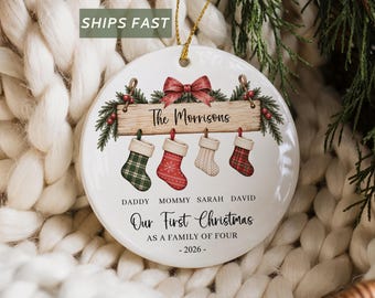 May include: A round white ceramic Christmas ornament featuring a wooden sign graphic with the text The Morrisons, four hanging stockings, and the text Daddy, Mommy, Sarah, David, Our First Christmas AS A FAMILY OF FOUR, and 2026.