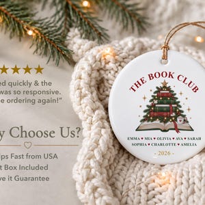 May include: A round white ceramic ornament featuring a graphic of a Christmas tree made of stacked books atop an open book. The text reads THE BOOK CLUB, followed by a list of names separated by hearts and the year 2026 at the bottom.