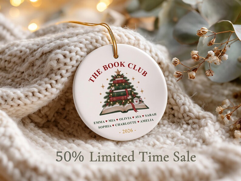 May include: A round white ceramic ornament featuring a graphic of a Christmas tree made of stacked books on an open book. The text reads THE BOOK CLUB, followed by a list of names separated by hearts, and the year 2026 at the bottom in gold.