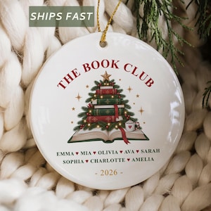 May include: A round white ceramic Christmas ornament featuring a graphic of a stack of books shaped like a tree, topped with a gold star. The text THE BOOK CLUB is printed in red above the tree, with a list of names and the year 2026 below.