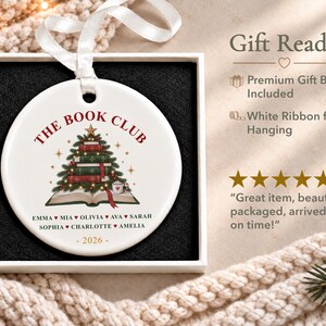 May include: A round white ceramic ornament featuring a graphic of a Christmas tree made of stacked books, with the text THE BOOK CLUB, a list of names including Emma, Mia, Olivia, Ava, Sarah, Sophia, Charlotte, Amelia, and the year 2026, with a white ribbon.