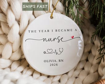 May include: A round white ceramic ornament featuring the black text THE YEAR I BECAME A nurse with a small red heart, a stethoscope and heart rate line graphic, and the personalized text OLIVIA, RN 2026, suspended by a thin gold string.