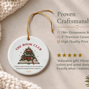 May include: A round white ceramic ornament featuring a gold hanging string. The design includes the text THE BOOK CLUB, a Christmas tree made of stacked books, and a list of names separated by hearts, with the year 2026 printed at the bottom.