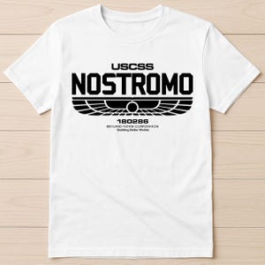 May include: White t-shirt featuring a black graphic. The design includes the text "USCSS NOSTROMO" above a stylized emblem and the text "180286" and "WEYLAND-YUTANI CORPORATION Building Better Worlds".