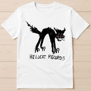 May include: White t-shirt featuring a black, stylized cat illustration with red eyes. The cat is arched and snarling, with the text "HELLCAT RECORDS" below the image. The design is a graphic print.
