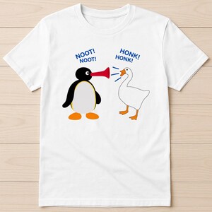 May include: White t-shirt featuring a cartoon penguin with a red horn and a white goose. The penguin says "NOOT! NOOT!" and the goose says "HONK! HONK!" in blue text. The shirt is a crew neck.