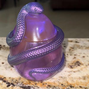May include: A translucent purple egg-shaped container with a removable lid, featuring a textured, iridescent purple snake coiled around the exterior. The surface of the container has a subtle glitter finish, and the snake has detailed scales.