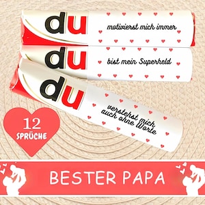 Puede incluir: Tres barras de chocolate blancas y rojas con texto en alem&aacute;n y detalles de corazones. El texto incluye frases como "motivierst mich immer" y "bist mein Superheld". Tambi&eacute;n se ve un coraz&oacute;n rojo con "12 Spr&uuml;che", junto con una pancarta que dice "BESTER PAPA".