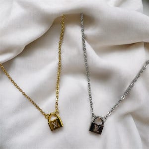 Puede incluir: Dos collares con colgantes de candado. Uno es dorado y el otro plateado. Ambos tienen una cadena delicada y un colgante de candado cuadrado con un logotipo. Los collares se exhiben sobre una superficie blanca texturizada.