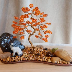 May include: A wire tree sculpture with orange beads, a white ceramic cat figurine, and brown stones on a brown base.