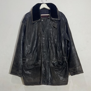 Puede incluir: Chaqueta de cuero negro vintage con cuello de piel sintética, cierre de botones y dos bolsillos delanteros. La chaqueta está colgada de una percha blanca sobre un fondo blanco liso. El cuero parece desgastado, dándole un aspecto clásico y envejecido.