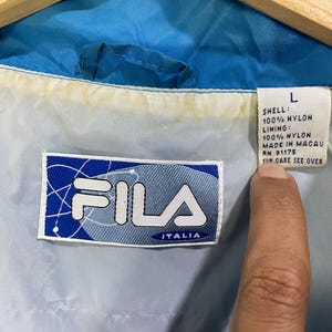 K&ouml;nnte beinhalten: Nahaufnahme eines blauen und wei&szlig;en Fila Italia Jackenetiketts. Das Etikett ist rechteckig und zeigt das Fila-Logo und das Wort "ITALIA" in Blau. Die Jacke ist aus Nylon und hat die Gr&ouml;&szlig;e L.