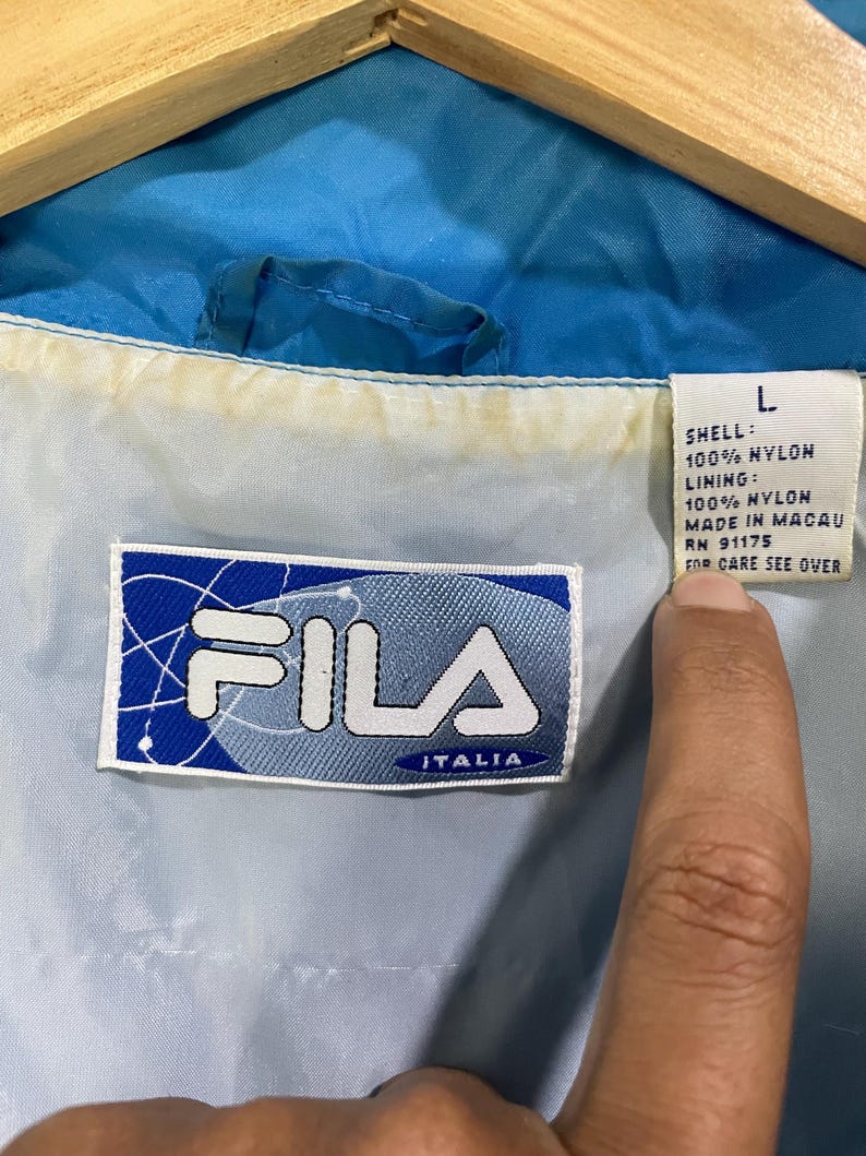 K&ouml;nnte beinhalten: Nahaufnahme eines blauen und wei&szlig;en Fila Italia Jackenetiketts. Das Etikett ist rechteckig und zeigt das Fila-Logo und das Wort "ITALIA" in Blau. Die Jacke ist aus Nylon und hat die Gr&ouml;&szlig;e L.