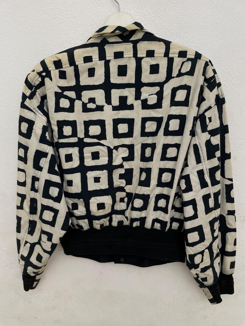 Może przedstawiać: Kurtka w stylu vintage z czarno-kremowym geometrycznym wzorem. Kurtka ma kołnierz, długie rękawy oraz czarny, prążkowany d&oacute;ł i mankiety. Wz&oacute;r składa się z czarnych kwadrat&oacute;w na kremowym tle.