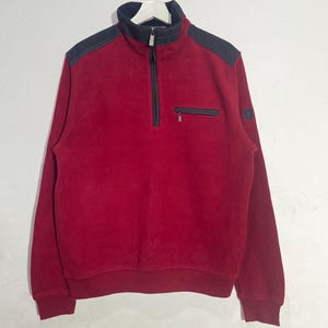 Op de afbeelding: Een rode fleece trui met een zwarte kraag en schouderaccenten. De trui heeft een kwart rits, een klein zakje en een logopatch op de mouw. Het kledingstuk hangt aan een witte hanger tegen een witte achtergrond.