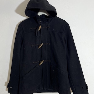May include: Black duffel coat with a hood. The coat has wooden toggle fastenings and two front pockets. The hood is attached and the coat has buttons on the sleeves. The coat is hanging on a white hanger.
